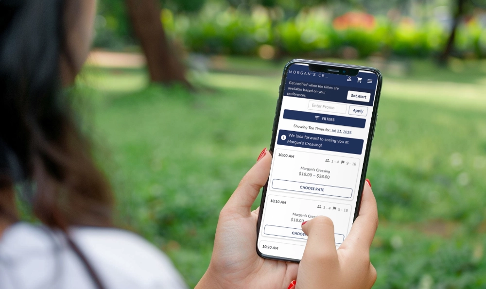 A woman holding a mobile phone open to the tee times booking form on the Morgan's Crossing website.